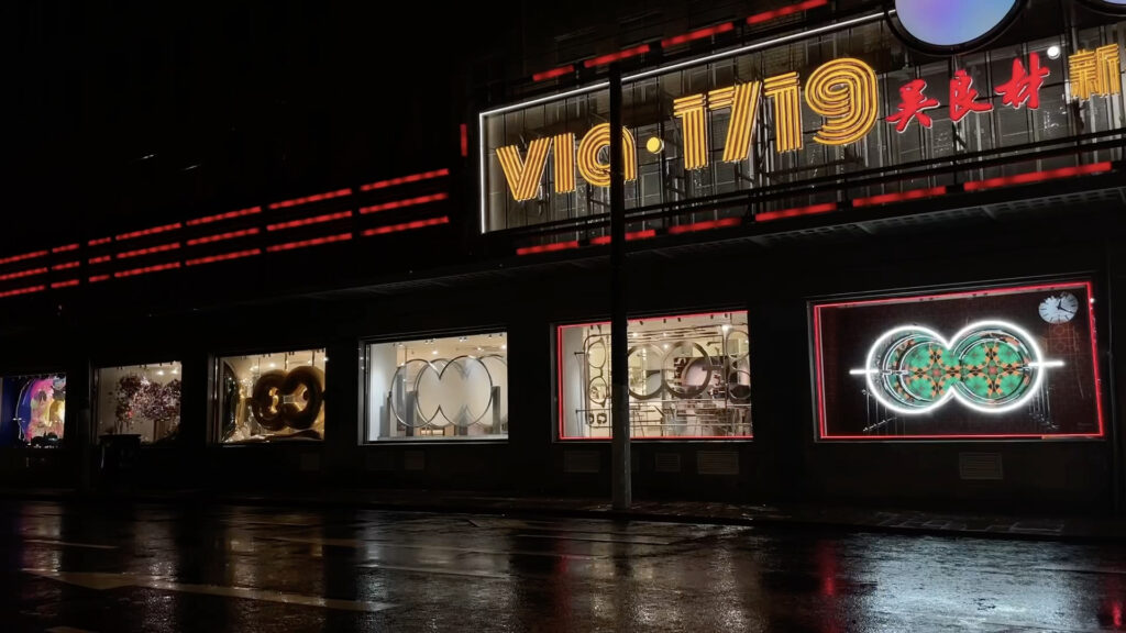 A row of brightly lit store windows and neon signs reflecting on a wet street at night, with VIC 1719 in large yellow letters above the storefront.