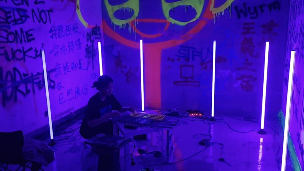 A person sits at a low table with electronic equipment in a room lit by four vertical purple neon lights. The walls are covered with colorful graffiti and a large, glowing face above the person.