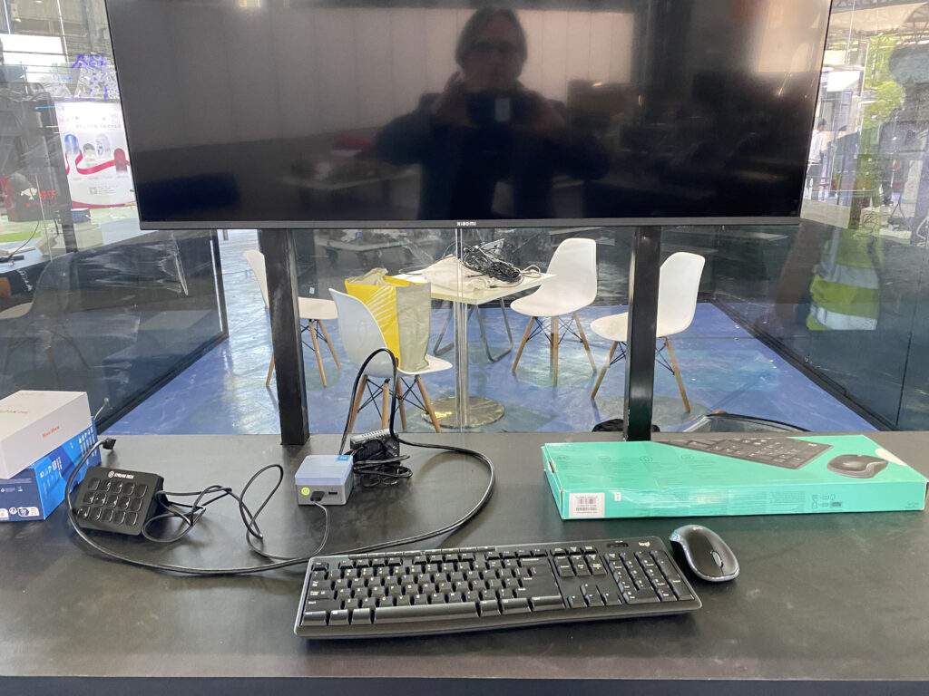 A computer setup with a keyboard, mouse, and monitor on a desk. Cables and a small device are connected. In the background are white chairs around a table in a glass-walled room. A person’s reflection is visible on the screen.