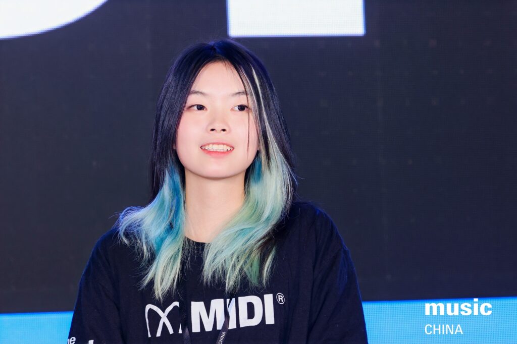 A young woman with black and teal hair smiles while standing in front of a dark background with the words music CHINA visible in the lower right corner. She is wearing a black MIDI t-shirt.