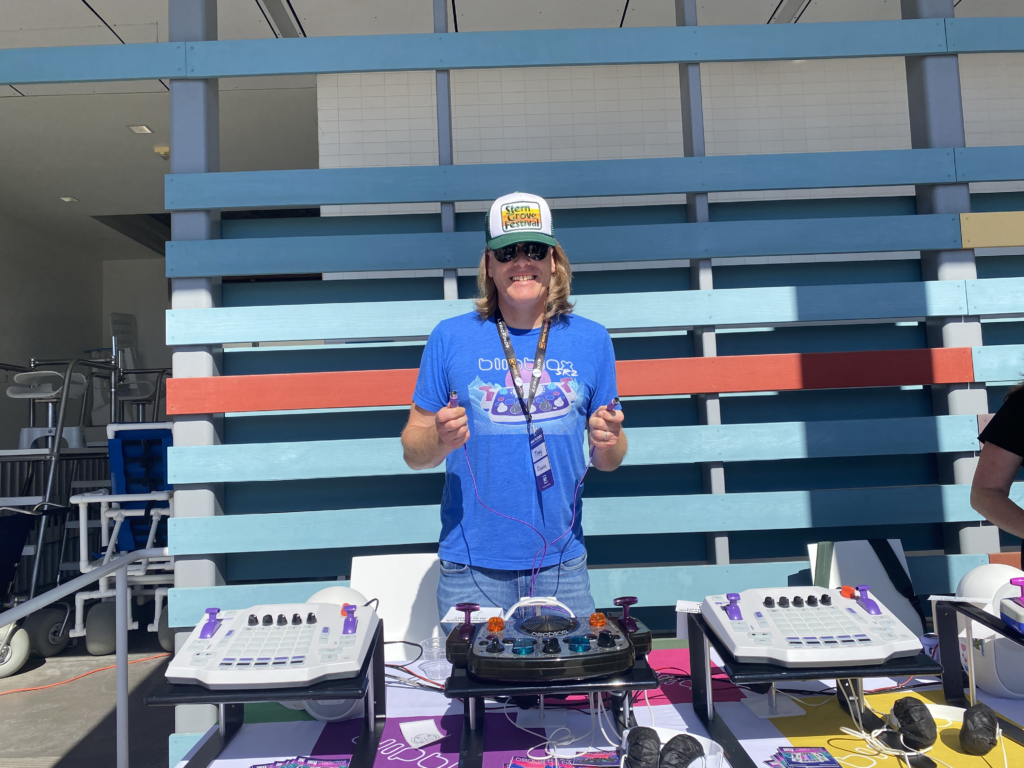A person wearing sunglasses and a hat stands smiling behind a table with electronic music equipment outdoors. Blue, red, and white horizontal panels form the background, and sunlight creates strong shadows.