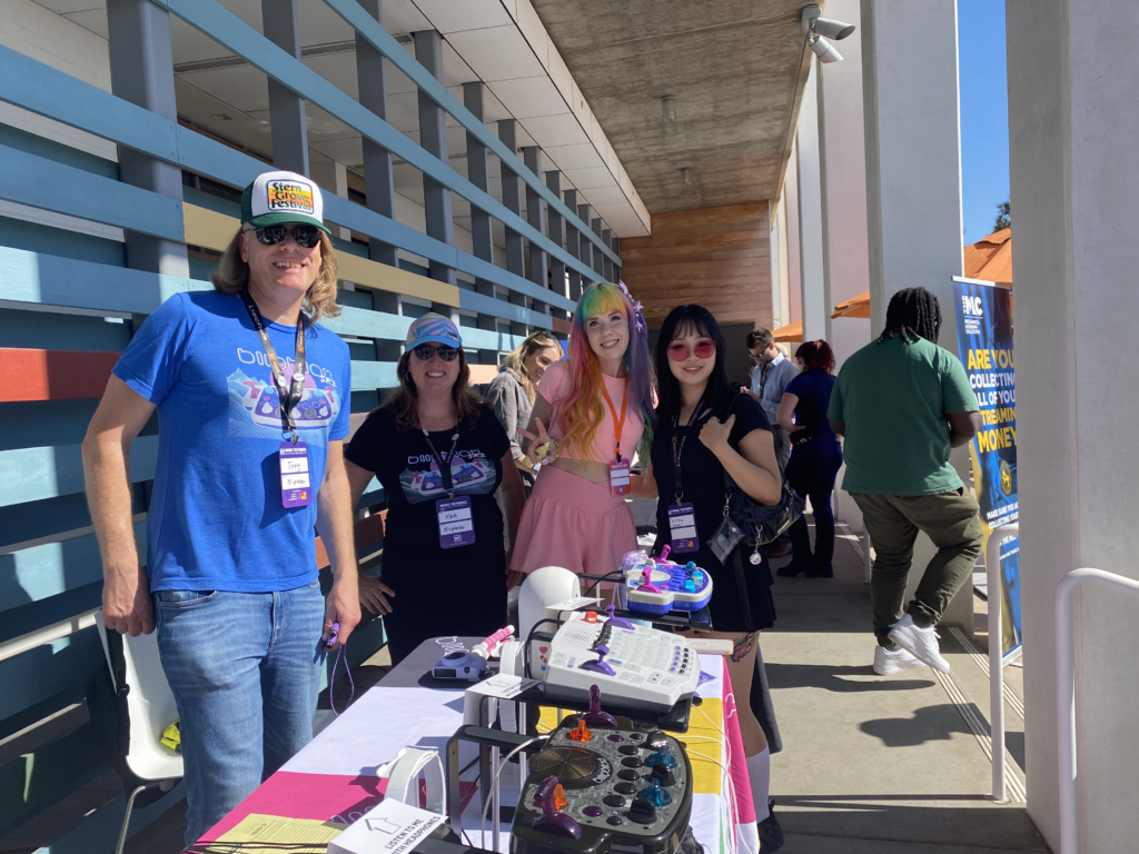 Four people stand smiling behind a table with electronic devices and crafts at an outdoor event. Two wear sunglasses, one has rainbow hair, and all wear event badges. Other attendees are visible in the background.