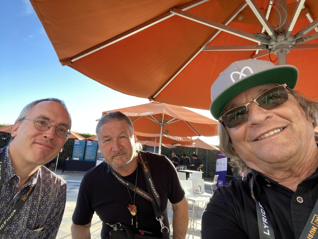 Three men pose for a selfie outdoors under large orange umbrellas. They wear conference lanyards and casual clothing. One man is smiling and wearing a gray cap. Clear blue sky is visible in the background.