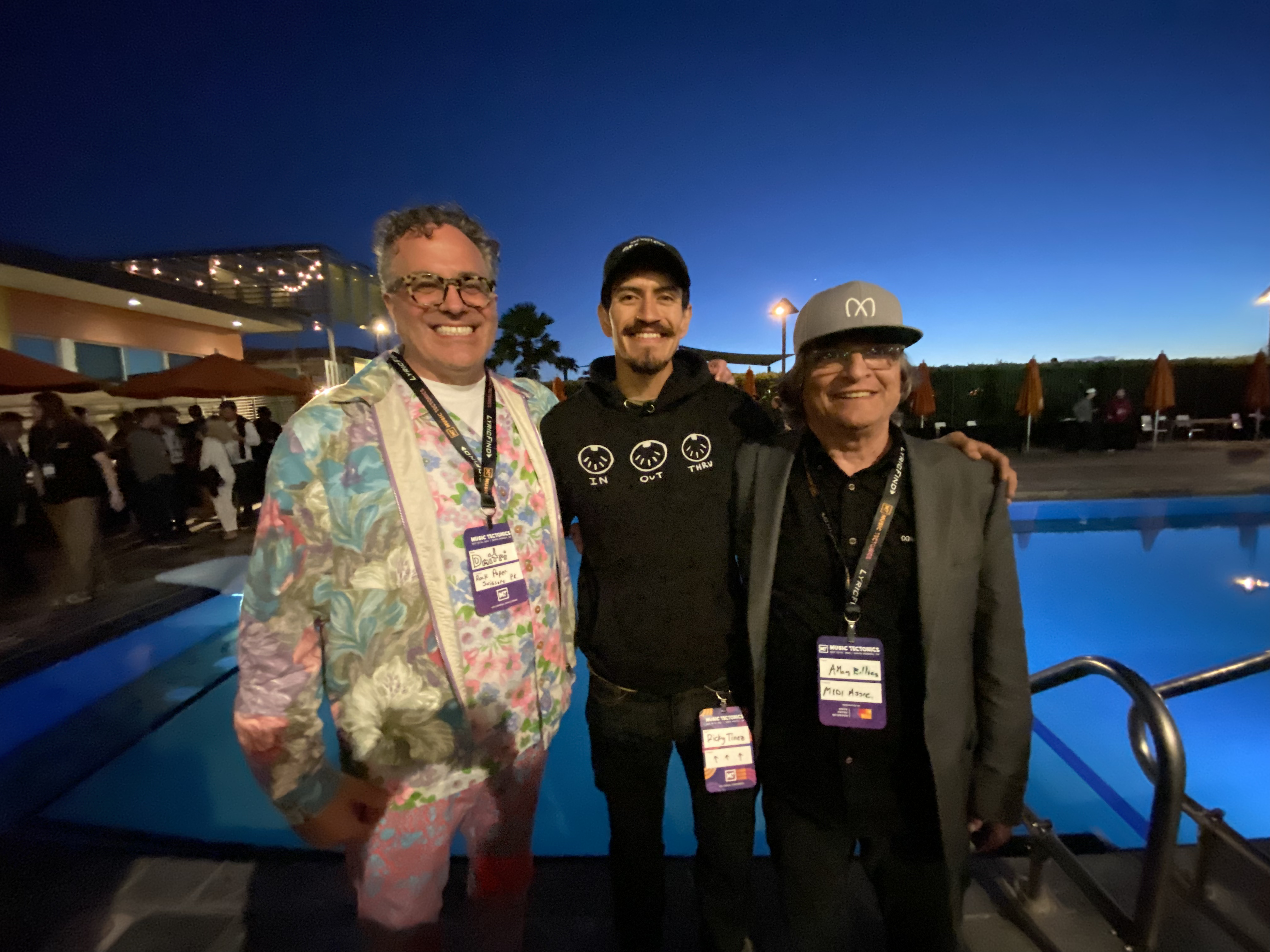 Three smiling people stand by a pool at dusk during an outdoor event, each wearing event badges. The sky is deep blue, and patio umbrellas and lights are visible in the background.