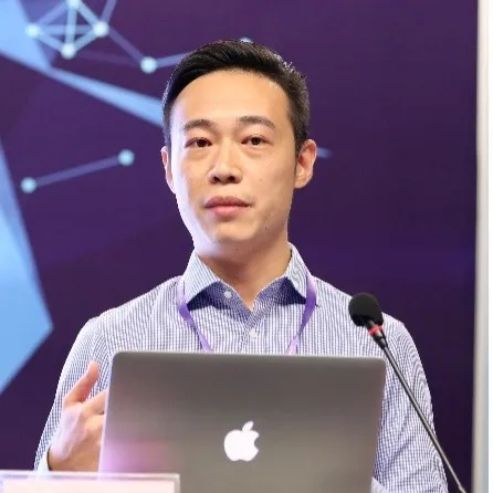 A man with short black hair and glasses wearing a black Kappa sweatshirt sits indoors, surrounded by audio equipment and instruments.