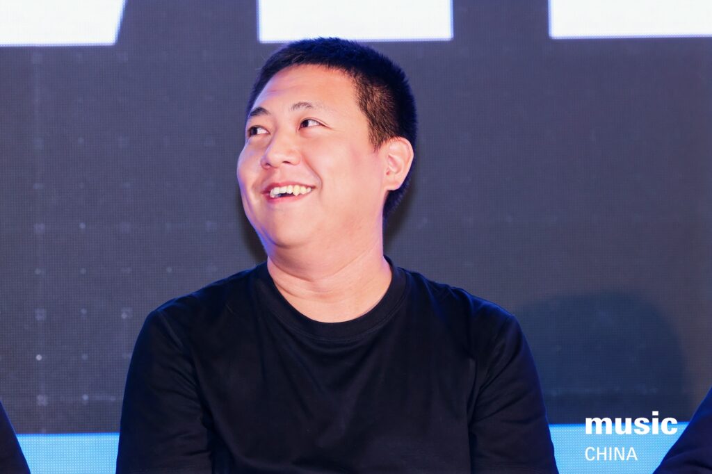 Six people sit onstage in front of a screen displaying headshots and text. One man stands and speaks, while the others sit and listen. The scene appears to be a panel discussion at a music event. The music CHINA logo is visible.