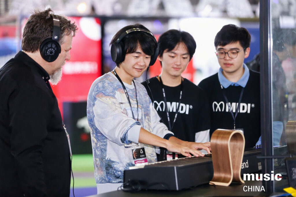 A young person plays an electronic keyboard while wearing headphones, observed by three others. They are at an indoor event, with music CHINA and MIDI logos visible in the background.