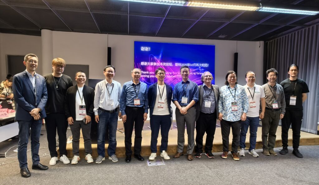 A group of twelve people stand in a row, smiling for a photo indoors. Behind them is a large screen with text in English and Chinese. They appear to be at a conference or event, with badges around their necks.