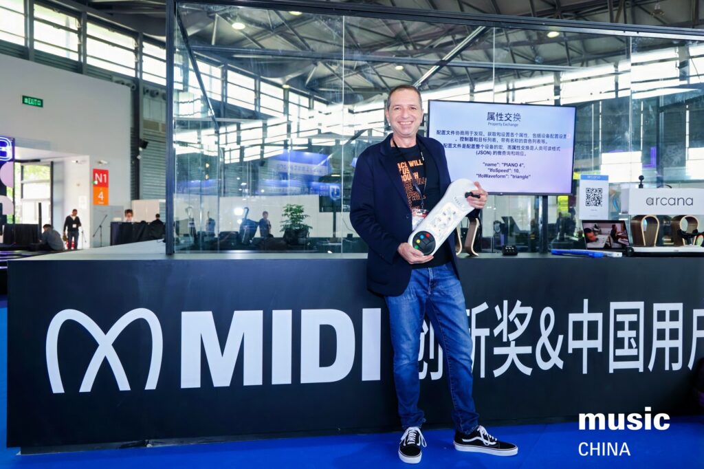A smiling man in casual clothes holds a white electronic keyboard in front of a MIDI display at a modern exhibition hall. The background features booths, signs, and screens. music CHINA is written on the blue floor.
