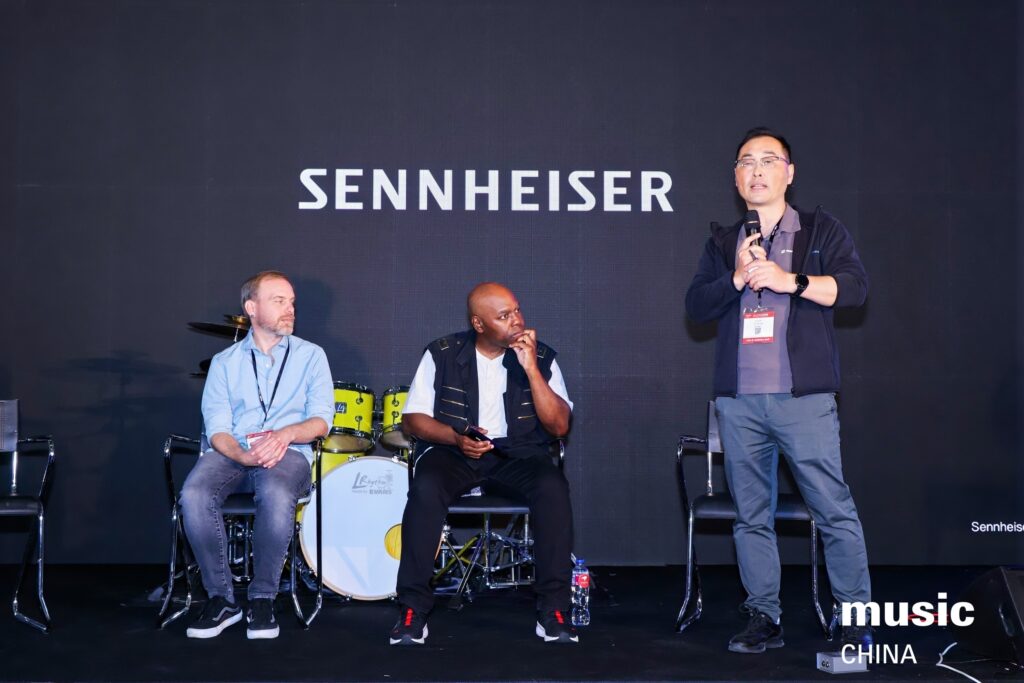 Three men are on a stage in front of a Sennheiser backdrop. Two are seated while one stands and speaks into a microphone. A drum set is visible behind them. The event is branded “music CHINA” in the corner.