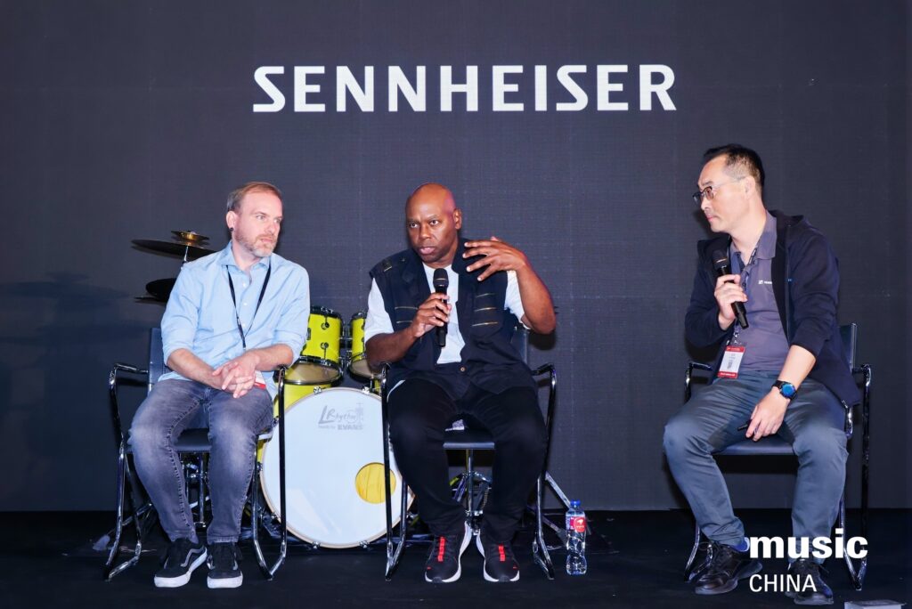 Three men sit on stage in front of a Sennheiser sign, one speaking into a microphone while the others listen. A drum set is behind them, and the Music China event logo appears in the corner.