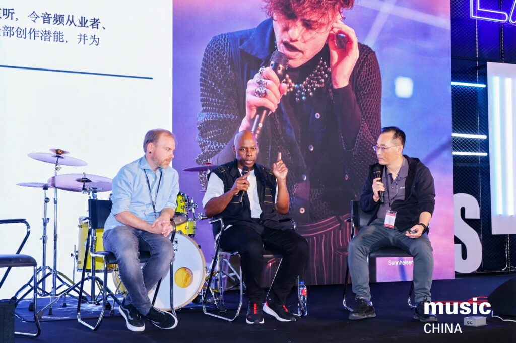 Three men sit on stage having a panel discussion at Music China, with drums behind them and a large photo of a singer performing projected in the background. One man speaks into a microphone while the others listen.