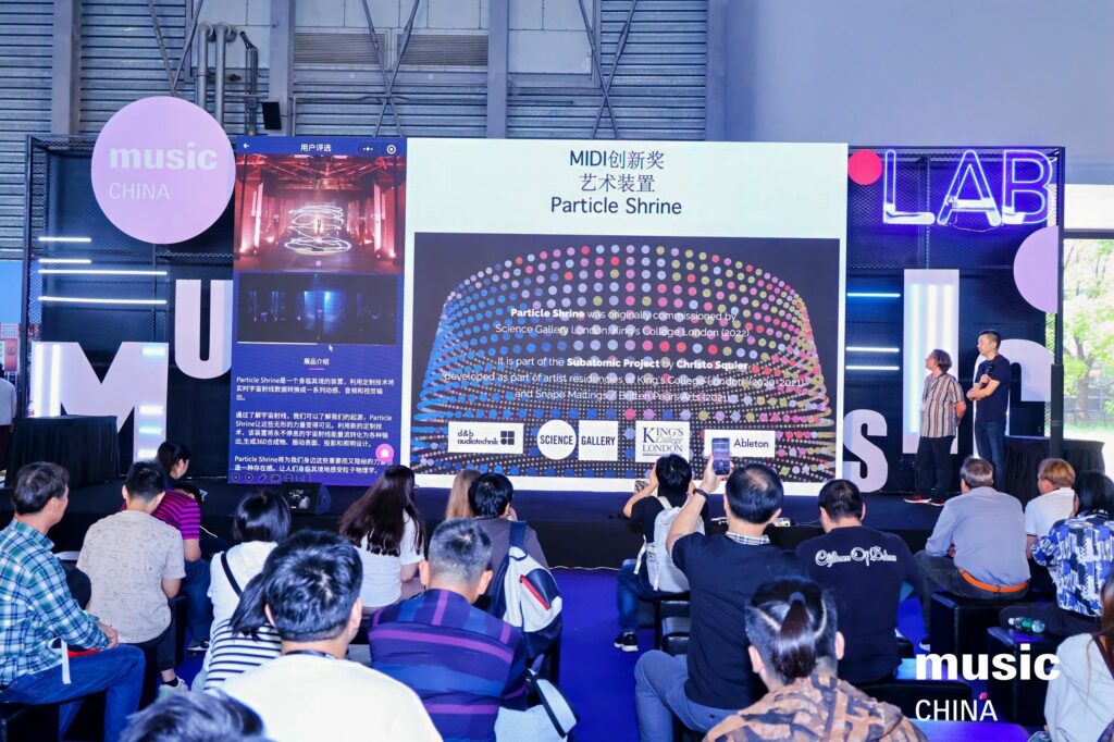 A seated audience watches a presentation on stage about Particle Shrine at Music China, with colorful visuals displayed on a screen and speakers standing to the side. The stage is decorated with large music and LAB signs.