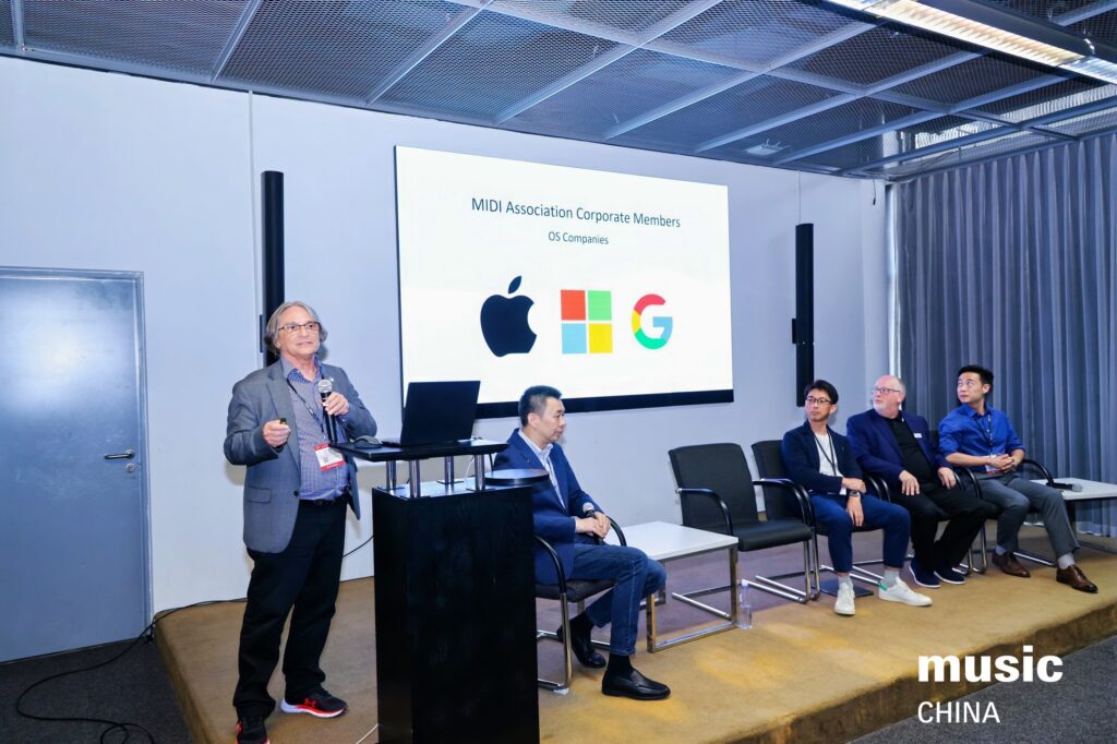 A man stands and speaks at a podium, presenting a slide showing the Apple, Google, and another logo, while four other men sit on a panel beside him at a music industry event in China.