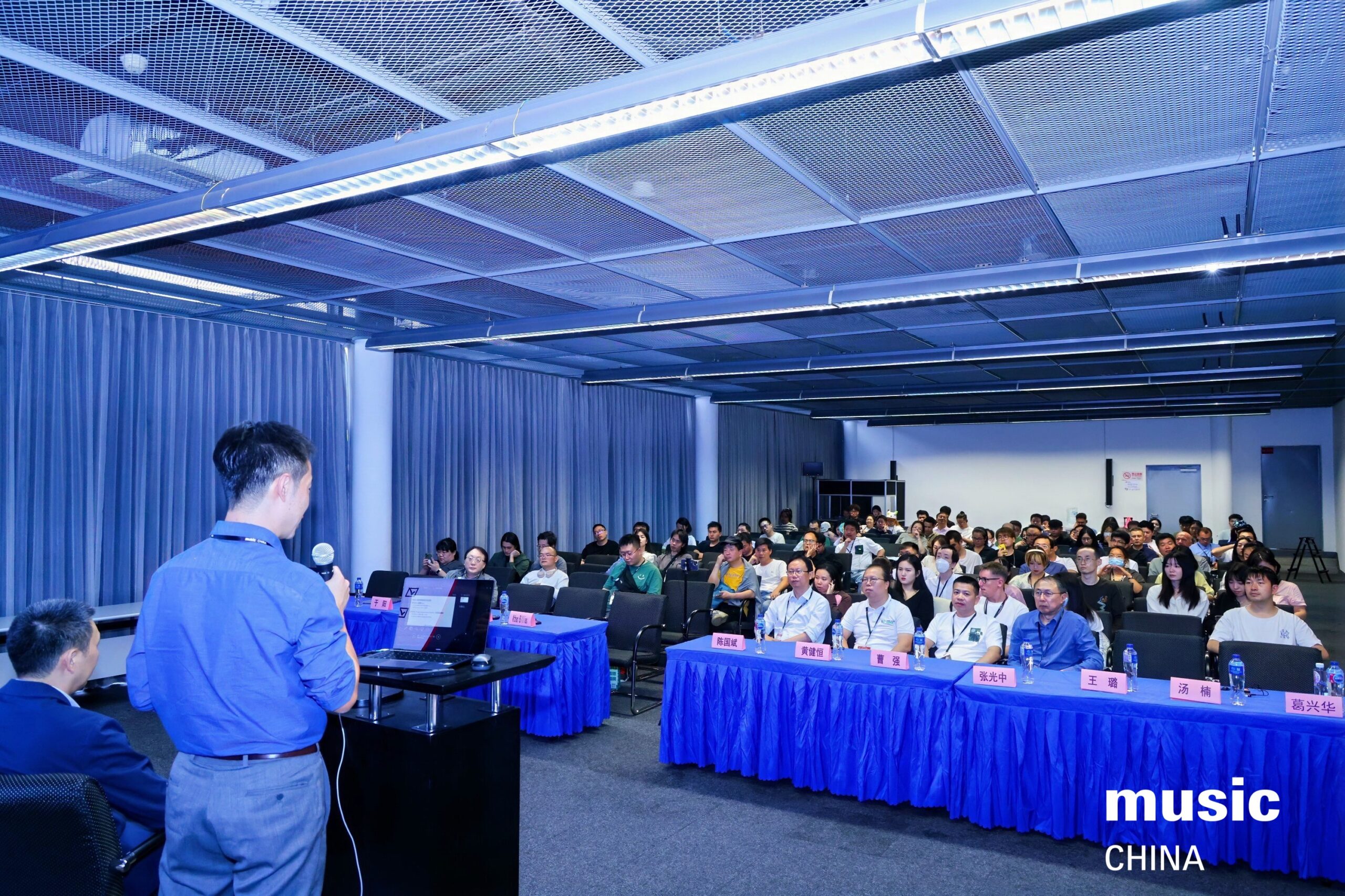 A speaker presents to a seated audience in a conference room with blue tablecloths. Nameplates are on the front table, and music CHINA is written in the bottom right corner.
