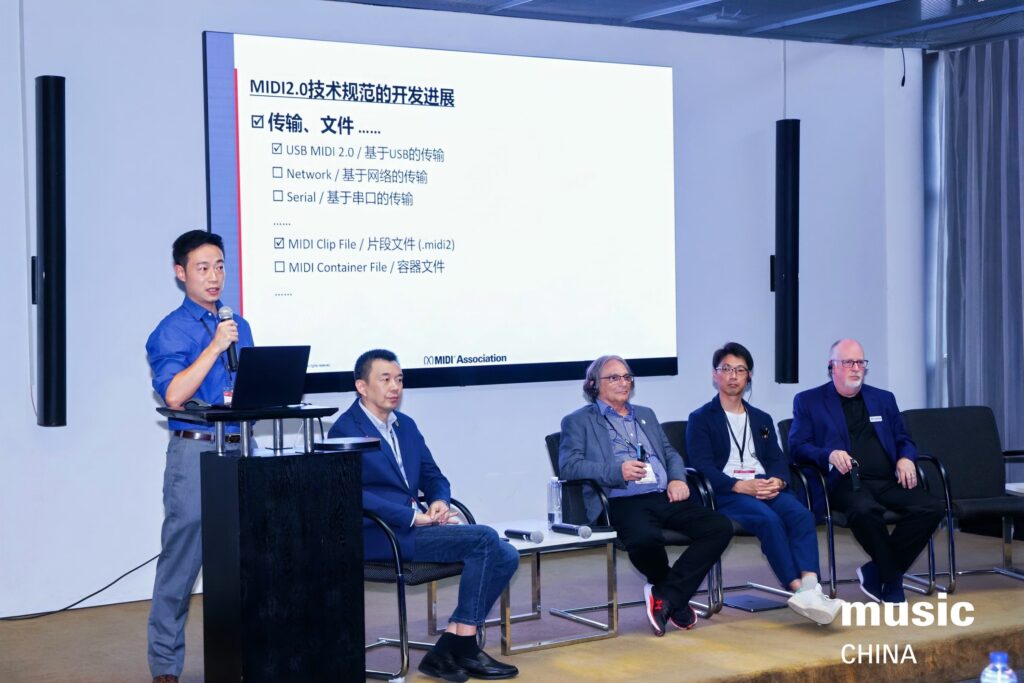 Four men sit on stage while another man stands at a podium speaking into a microphone. Behind them is a large screen displaying information about MIDI 2.0 data transmission in Chinese and English. The event is labeled music CHINA.