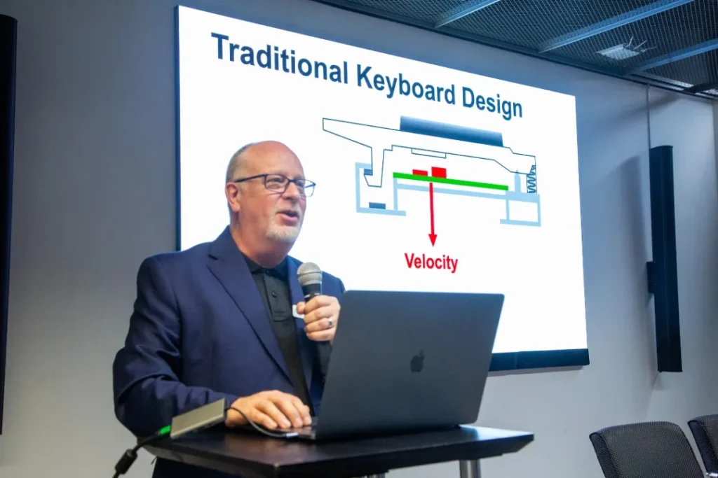 A man in glasses and a dark blazer speaks into a microphone while standing at a podium with a laptop. Behind him, a screen displays a diagram titled “Traditional Keyboard Design” with an arrow labeled “Velocity.”.