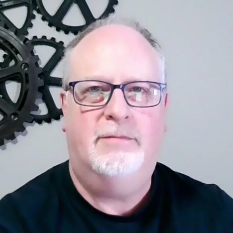 A middle-aged man with short gray hair, glasses, and a closely trimmed white beard is looking at the camera. He is wearing a dark shirt, with large black gear decorations on the light gray wall behind him.