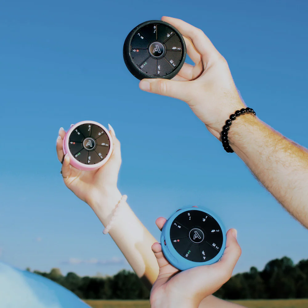 Three hands hold round, black and blue electronic devices with buttons and symbols against a clear blue sky, with trees in the background. Each hand wears a different bracelet.