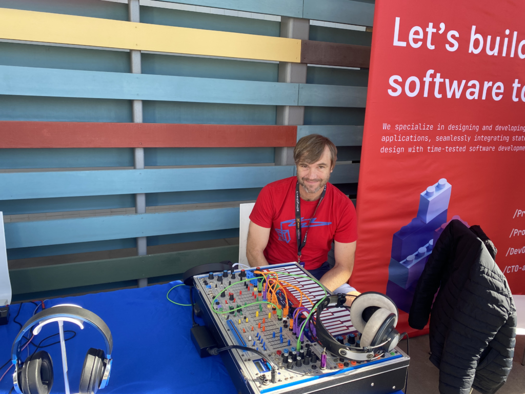 A man sits at a table with audio equipment and headphones outside, next to a red banner that partially reads, “Let’s build software t...”. The background features colorful horizontal wooden slats.