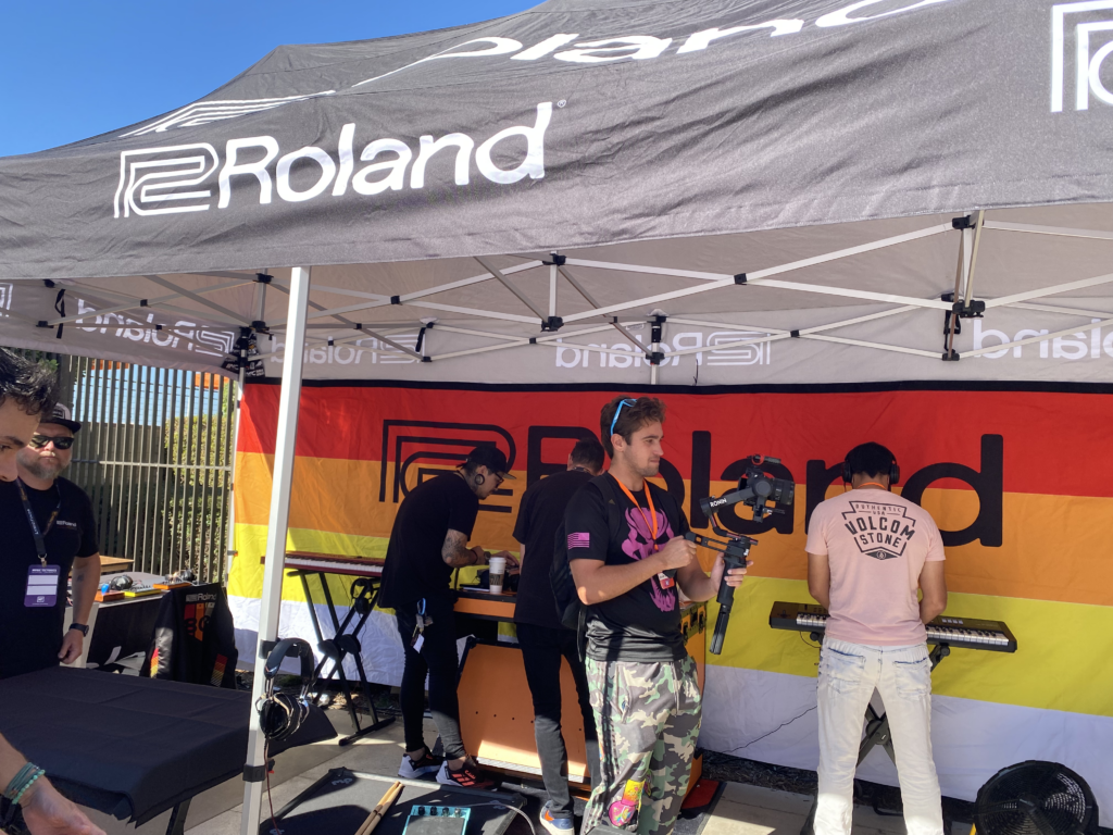 People are playing keyboards and synthesizers under a black Roland tent at an outdoor event. A colorful Roland banner is displayed behind them, and the scene is lively and sunny.