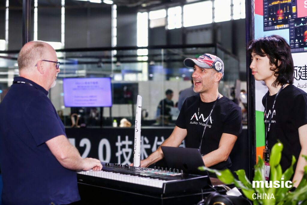 Three people interact at a music technology booth; one man plays a keyboard while another man, wearing a cap and smiling, and a woman stand nearby. Music equipment and screens are visible in the background. Text reads “music CHINA.”.