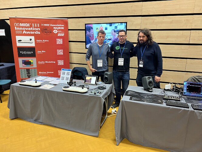 Three men stand behind a booth at a technology event, showcasing audio equipment, speakers, and various electronic devices. A red sign with MIDI Innovation Awards and QR codes is displayed on the table.