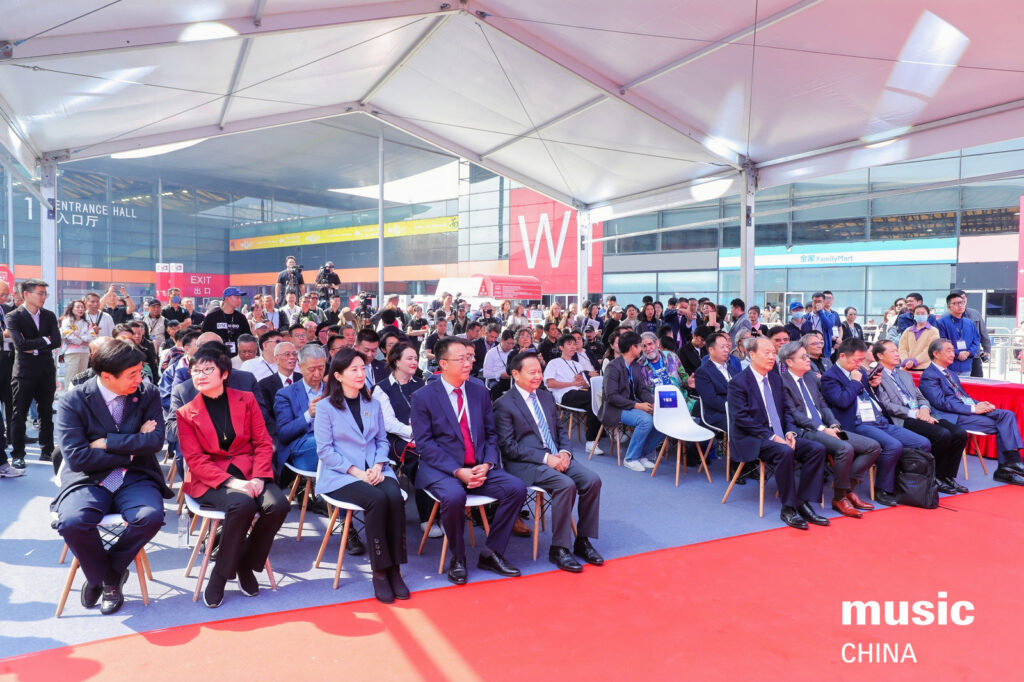 A large group of people in formal attire sit on chairs under a white tent at an outdoor event. There is a red carpet, and some audience members are watching attentively. The background shows buildings with large glass windows.