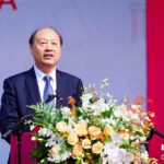 A man in a dark suit speaks at a podium decorated with yellow and white flowers, with “music CHINA” written in white on a red background behind him.