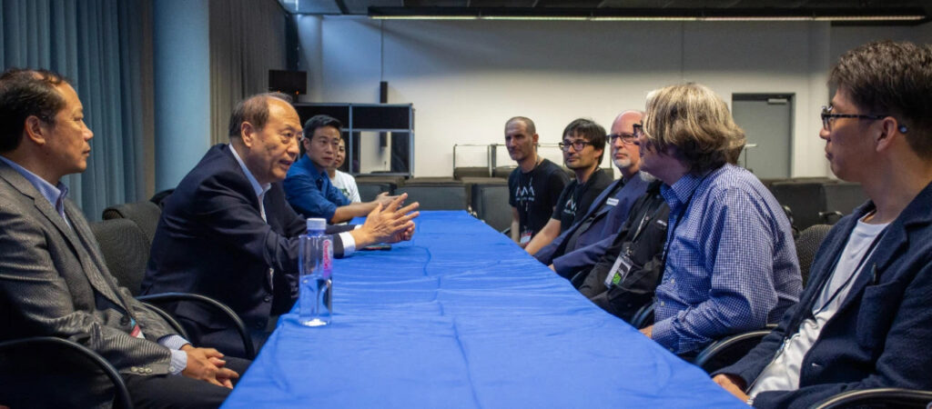Eight men sit on opposite sides of a long table covered with a blue cloth, engaged in a discussion in a conference room setting. The atmosphere appears serious and professional.