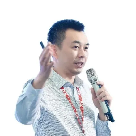 A man wearing a light-colored shirt and red lanyard is speaking into a microphone and gesturing with his other hand, with a white background behind him.