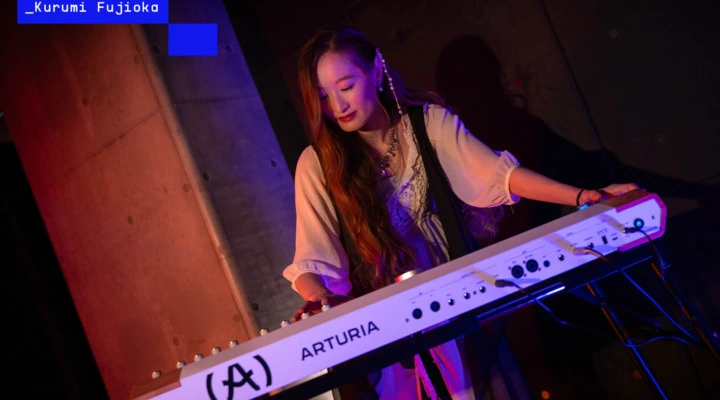A woman in a white dress plays an Arturia keyboard on stage under colorful lighting, with long hair draped over her shoulder and focused expression. The name _Kurumi Fujioka appears in the upper left corner.