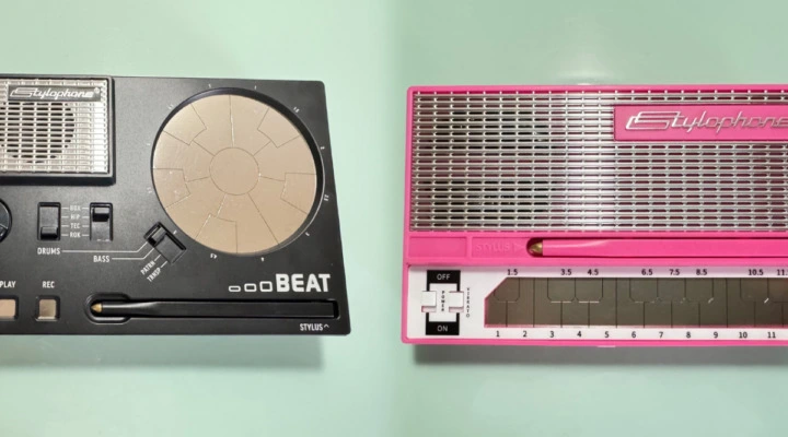 Two Stylophone instruments are shown on a light green background: one black and silver with circular controls labeled BEAT, and one pink and silver with rectangular keys and an on/off switch.
