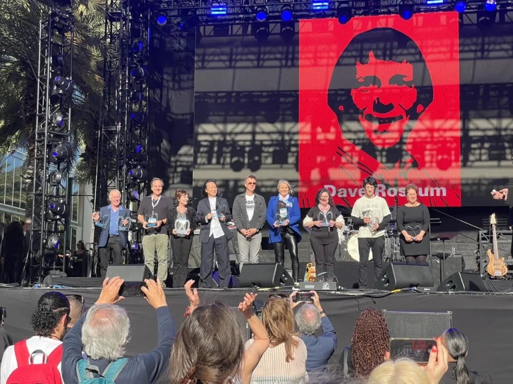 A group of people stand on an outdoor stage holding awards, while an audience takes photos. Behind them is a large red and black illustration of a smiling face and the name Dave Rosum. Palm trees are visible on the left.
