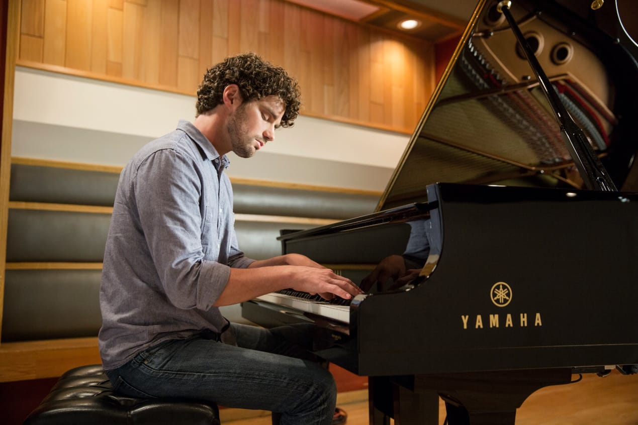 A man with curly hair and a casual shirt plays a Yamaha grand piano in a modern studio with wooden walls and padded benches.
