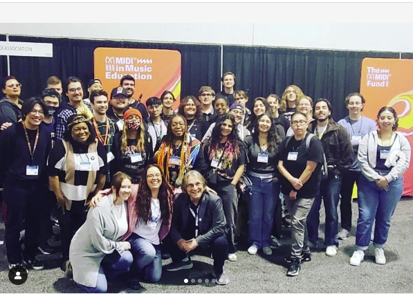 A large group of people pose and smile together indoors in front of colorful banners that read “MIDI in Music Education” and “The MIDI Fund.” Most are wearing name badges.