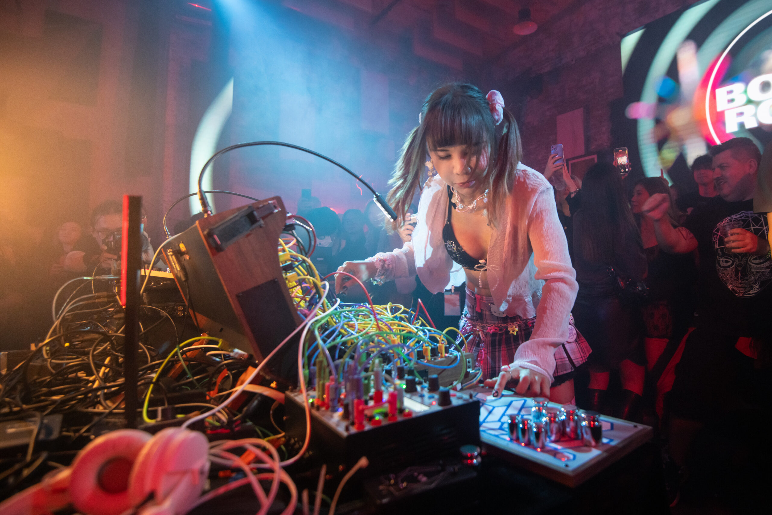 A DJ with pigtails performs live, surrounded by synthesizers and cables, as a crowd of people watch and dance under colorful lights in a vibrant club setting.