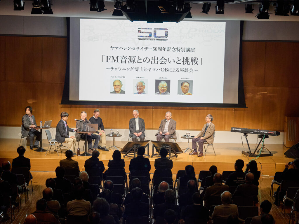 A panel of seven people sit on stage in front of an audience, with a large screen behind them displaying Japanese text, photos of four individuals, and a logo reading 50. A keyboard is visible on the right side of the stage.