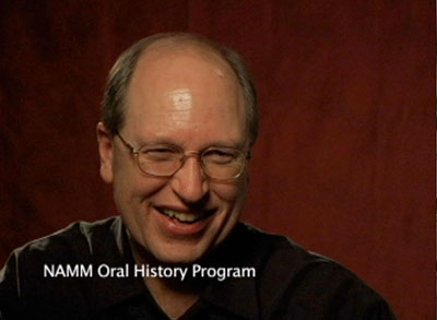 A smiling man with glasses sits in front of a dark red background. The text “NAMM Oral History Program” appears on the lower left corner of the image.
