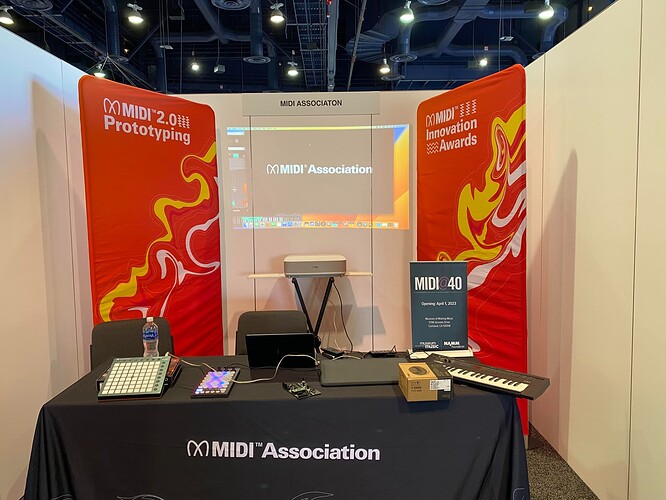 A booth at an event features two red banners for MIDI Association, a table with electronic music equipment, a keyboard, a laptop, and a sign promoting MIDI 2.0 Prototyping and Innovation Awards. A screen displays the MIDI Association logo.