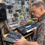A person adjusts knobs on an audio mixer and plays a synthesizer keyboard, surrounded by a large modular synthesizer setup with many colorful cables and components.