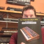 A person holding a book titled The Prophet from Silicon Valley: The Complete Story of Sequential Circuits in front of their face, with shelves of synthesizers in the background.