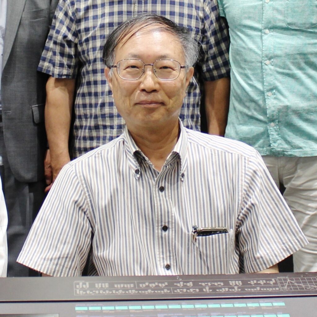 A man with glasses and gray hair, wearing a striped short-sleeve shirt with a pen in the pocket, sits in front of an electronic control panel. Others stand behind him, mostly out of frame.