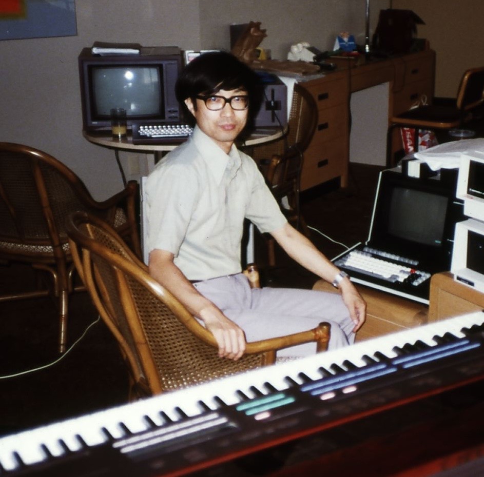 A person wearing glasses and a light-colored shirt sits on a wicker chair in a room with vintage computer equipment and a piano keyboard in the foreground.