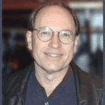 A middle-aged man with glasses and short hair is smiling at the camera. He is wearing a dark shirt and a black leather jacket, with an out-of-focus background behind him.
