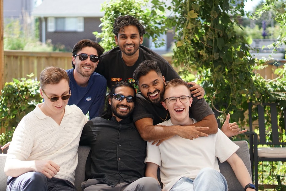 Six men sit closely together on an outdoor sofa, smiling and laughing. The group appears happy and relaxed, with greenery and wooden fencing in the background. Some are wearing sunglasses.