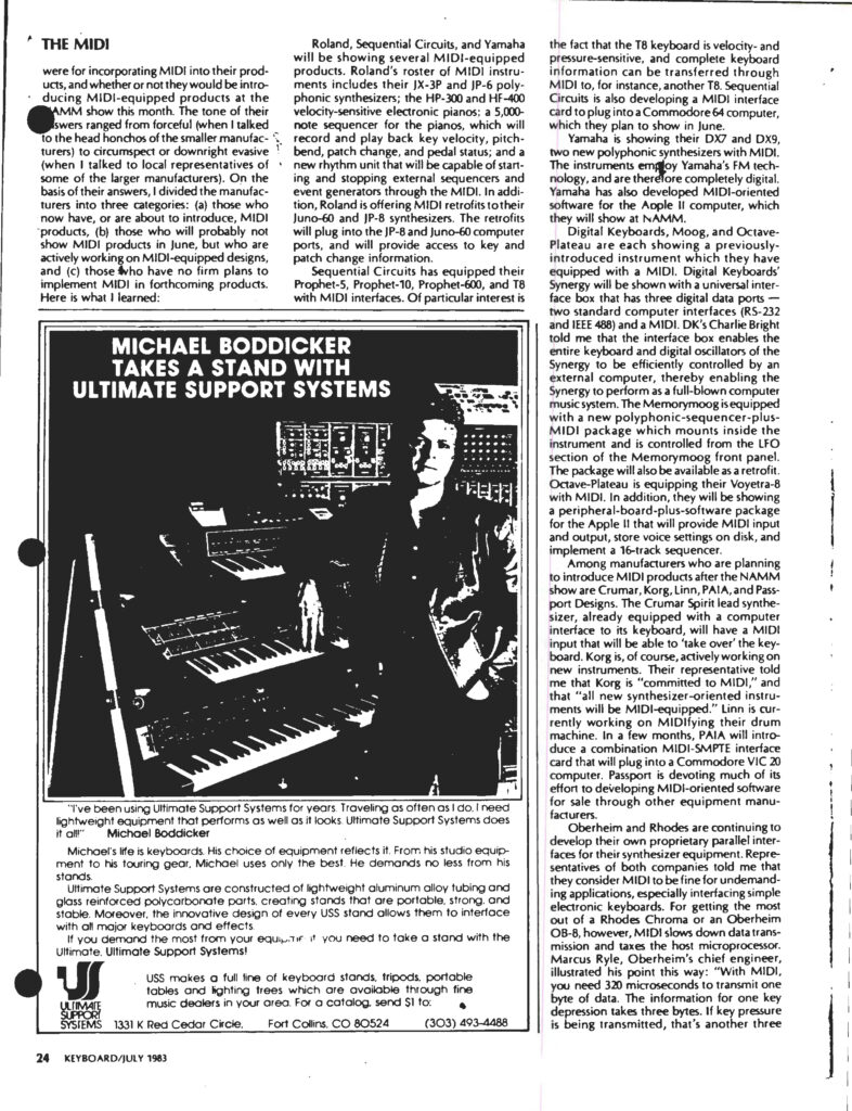 A man sits at a desk surrounded by keyboards, synthesizers, and audio equipment in a professional music studio. He looks at the camera, holding a pen, with audio cables and mixers visible on the desk.