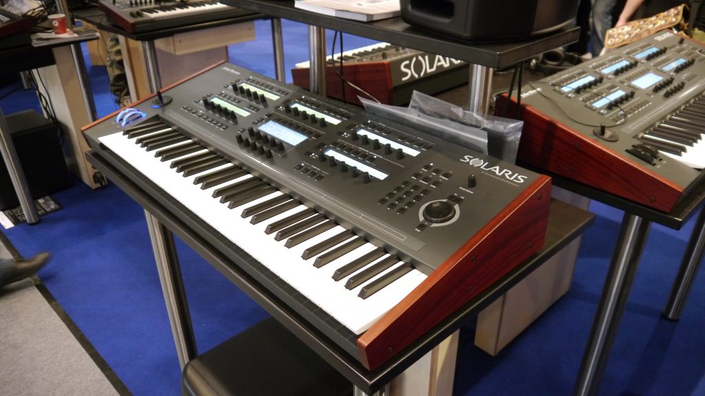 A black Solaris synthesizer keyboard with wooden side panels is displayed on a stand at an exhibition, surrounded by other musical equipment on a blue carpeted floor.