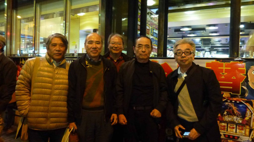 Five men stand together and smile for a group photo at night in front of a brightly lit building with large windows and colorful signs. They are wearing jackets and glasses.