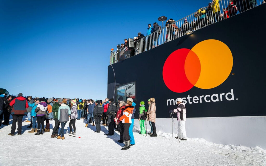 People in winter clothing gather outside on snow near a large black structure displaying the Mastercard logo. More people are seen standing on an upper level under a clear blue sky.